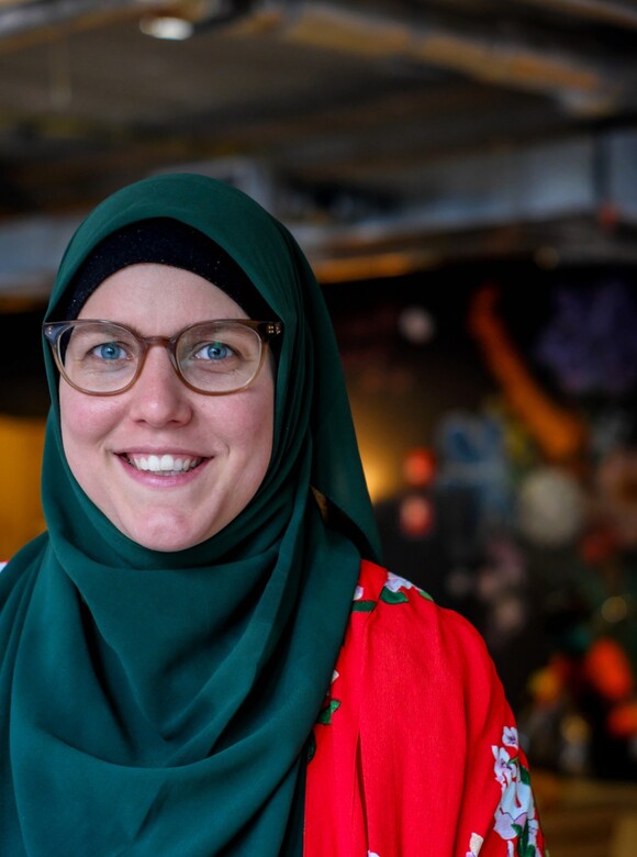 Foto van Sari Noordwest. Sari is een witte vrouw en draagt een donkergroene hijab. Ze heeft lichtblauwe ogen en draagt ook een bril. Verder heeft ze een roodkleurige trui aan.