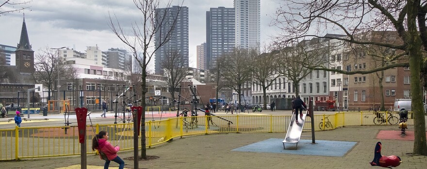 Een groep kinderen speelt op een grote speeltuin. Op de achtergrond is de skyline van Rotterdam te zien.