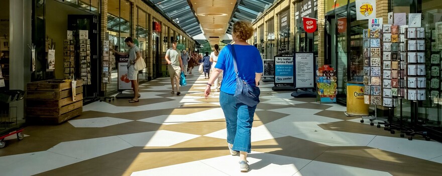 Witte vrouw met blond haar loopt in overdekte winkelstraat, met haar rug naar de kijker toe 