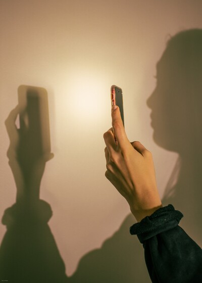 Een hand houdt een smartphone vast en maakt een foto. Op de muur erachter is de schaduw van een gezicht te zien.