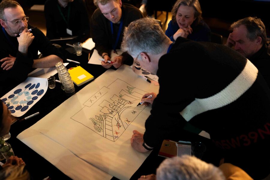 Een groep mensen aan een tafel praat met elkaar terwijl één persoon iets tekent op het grote vel in het midden van de tafel