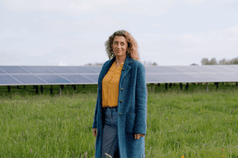 Een vrouw staat in het midden van een groot grasveld en achter haar is een veld met zonnepanelen te zien