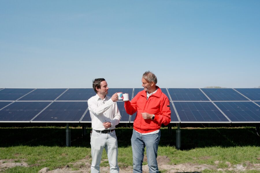 Twee mannen staan voor een veld met zonnepanelen en proosten met mokken