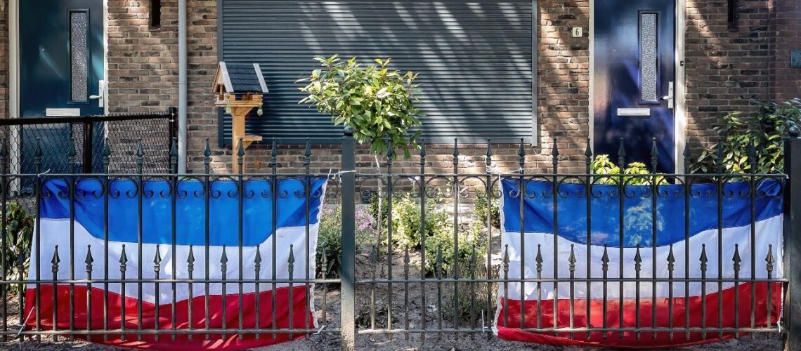 Het raam aan de voorkant van een huis is met een rolluik verborgen en langs het hek van de voortuin zijn twee Nederlandse vlaggen gehangen