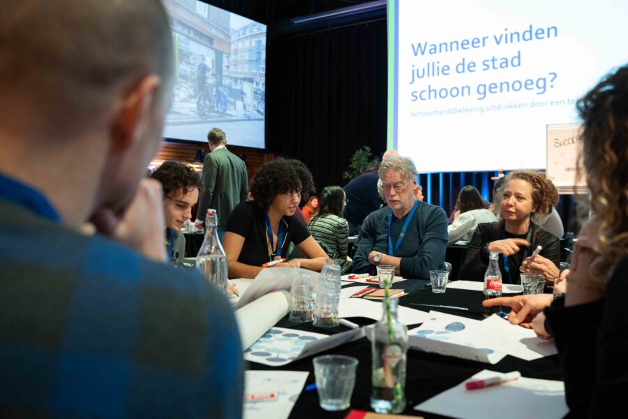 Een groep mensen zit aan een tafel met een aantal papieren erop en praat met elkaar. Op de achtergrond is een presentatie te zien waar op staat: 'Wanneer vinden jullie de stad schoon genoeg?'