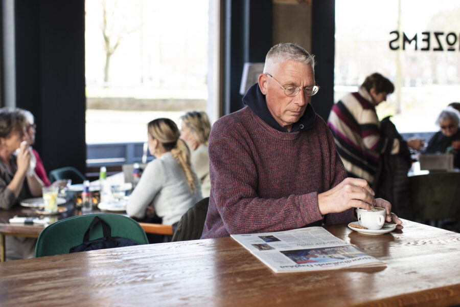 Een man zit aan een tafel in een café en roert in zijn kopje koffie terwijl hij de krant leest.