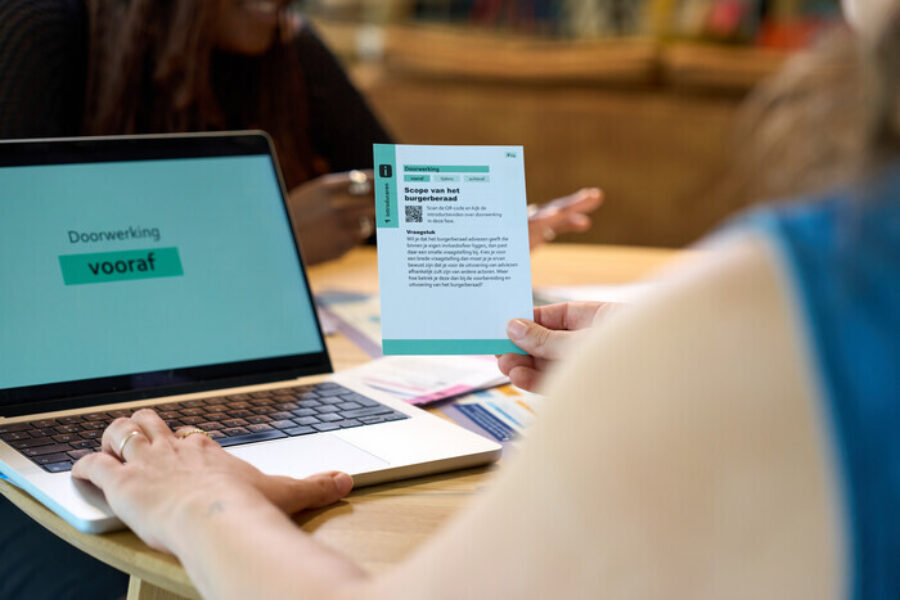 Iemand zit achter de laptop en houdt een kaart omhoog waarop staat: scope van het burgerberaad.