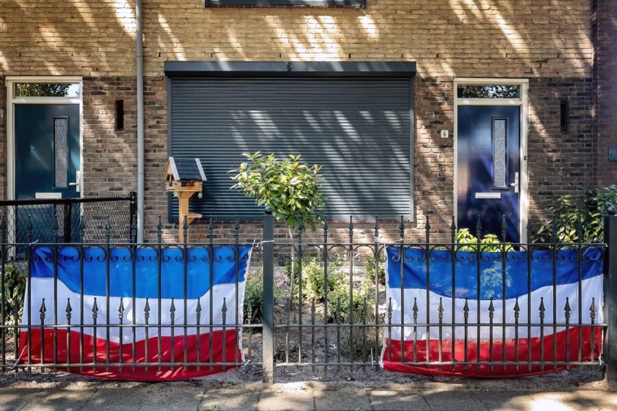 Het raam aan de voorkant van een huis is met een rolluik verborgen en langs het hek van de voortuin zijn twee Nederlandse vlaggen gehangen